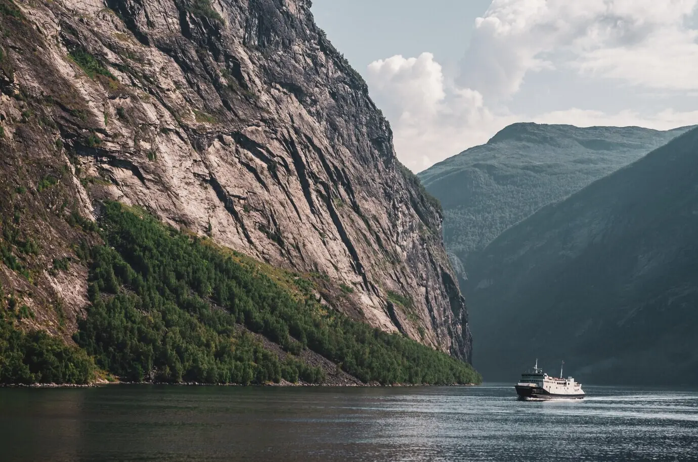A lone ship on a lake surrounded by tall rocky mountains beneath a cloudy sky in Norway.